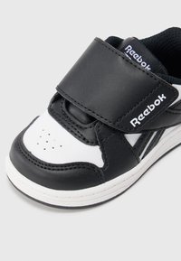 Melnas un baltas sporta kedas ar gludu ādas augšdaļu, Velcro siksnu, perforētu pirkstu daļu ventilācijai un gumijas zoli ar baltu šuvju detaļām.