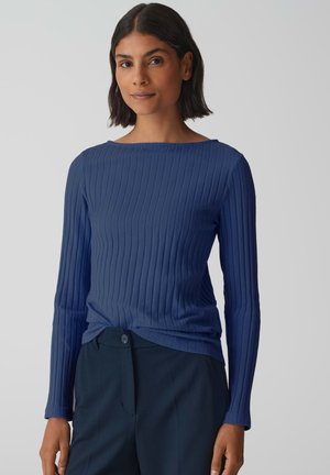 Blaues, geripptes Langarmshirt mit Bootsausschnitt, aus strukturiertem Stoff und figurbetonter Silhouette. Kombiniert mit einer dunklen Hose.