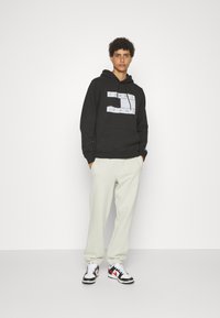 Felpa nera con logo "TOMMY JEANS" in bianco, abbinata a pantaloni sportivi beige chiaro e sneaker nere con dettagli rossi.