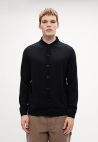 Μαύρο πλεκτό κασκόλ με μυτερές γιακά, μακριά μανίκια, κλείσιμο μπροστά με κουμπιά και ριγέ τελείωμα. Λείο υλικό, μινιμαλιστικός σχεδιασμός.