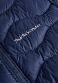 Marineblau gesteppte Jacke aus glänzendem synthetischen Material, verfügt über ein graues "PeakPerformance"-Logo, einen Reißverschluss und dezente Steppmuster.