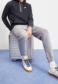 Grå sweatpants och en svart hoodie med en vit logotyp, kombinerat med vita högre sneakers, sittande på en silverfärgad resväska med hjul.