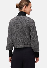 Femme avec les cheveux en chignon portant un short taille haute noir et une veste cropped grise à motifs sur un col roulé noir, souriant de côté.