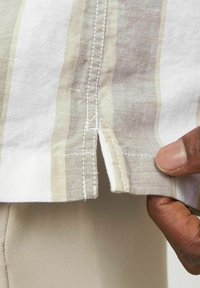 Gestreepte linnen blouse in beige en wit, met een zijsplit en gestikte zoom. Close-up toont de stofstructuur en interactie met de hand.