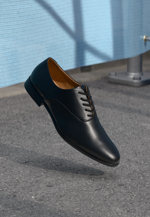Scarpa da uomo in pelle nera sopra una superficie grigia texturizzata con sfondo di parete blu e ombre di finestra.
