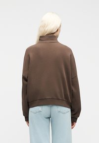 Άτομο με μακριά ξανθά μαλλιά, φοράει καφέ φούτερ με ζιβάγκο και ανοιχτό μπλε τζιν, στραμμένο πλάτη σε απλό φόντο.