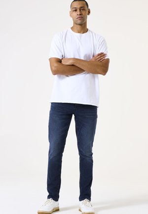 Camiseta blanca de algodón, jeans de mezclilla azules y zapatillas blancas con detalles en marrón. Modelo de pie con los brazos cruzados, fondo neutro.