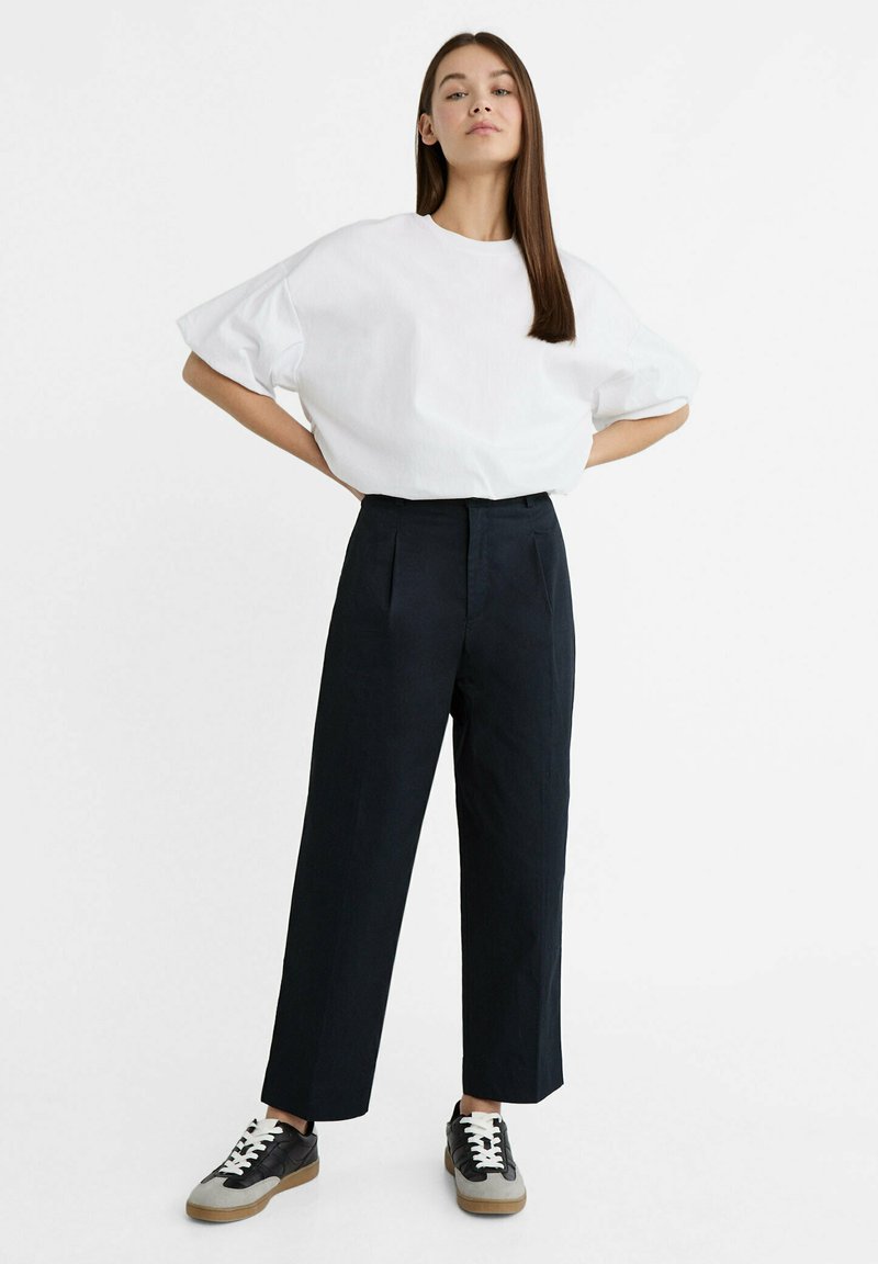 T-shirt oversize bianco in cotone, pantaloni neri a vita alta con pieghe e sneakers nere con lacci bianchi e suole in gomma.