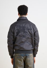 Black bomber jacket with embroidered "Flying Tigers" design, ribbed cuffs and hem, glossy nylon material, worn with light denim jeans.