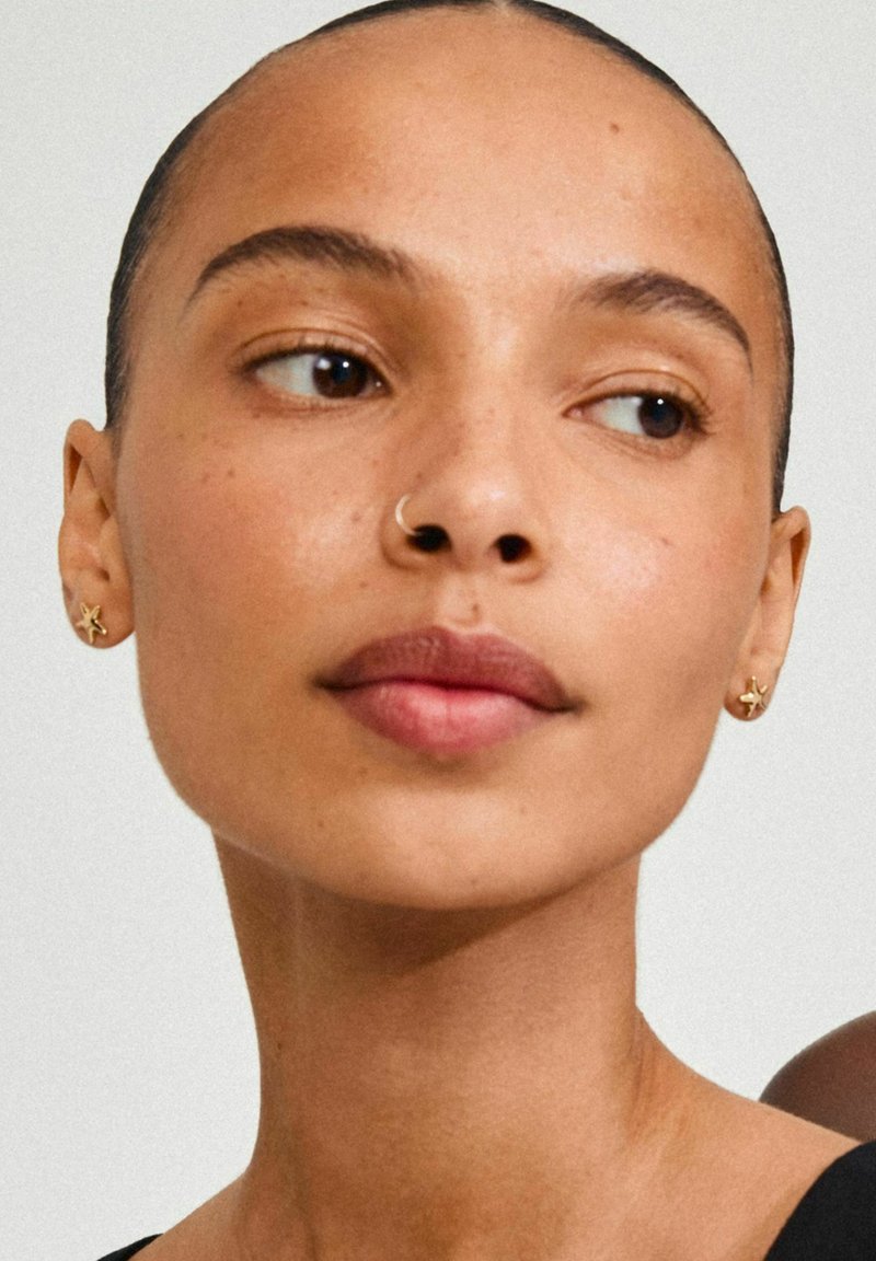 Gouden stervormige oorbellen en een neusring passen mooi bij de gladde teint van het onderwerp en de natuurlijke make-up, met kort haar en een neutrale achtergrond.