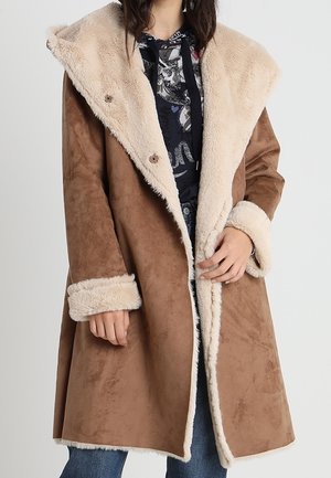 Długi brązowy płaszcz zamszowy z szerokim kremowym kołnierzem i mankietami z sztucznego futra, noszony na czarnej bluzie z kapturem i niebieskich dżinsach.