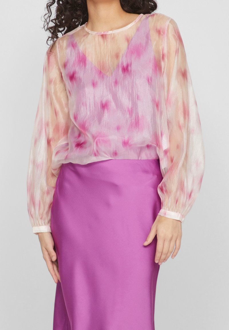 Femme portant un chemisier transparent à manches longues avec des motifs roses et rouges sur un caraco rose, associé à une jupe violette brillante.
