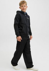 Čierna mikina na zips s prednými vreckami, skombinovaná s voľnými čiernymi cargo nohavicami. Model má na sebe biele tenisky a pod nimi kockovanú košeľu.