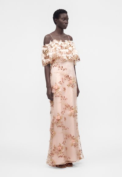 Γυναίκα που φοράει αμπιγιέ φόρεμα μήκους μέχρι το πάτωμα σε ροζ απόχρωση, με λουλουδάτες εφαρμογές στους ώμους και λεπτές τιράντες, στέκεται μπροστά σε απλό φόντο.