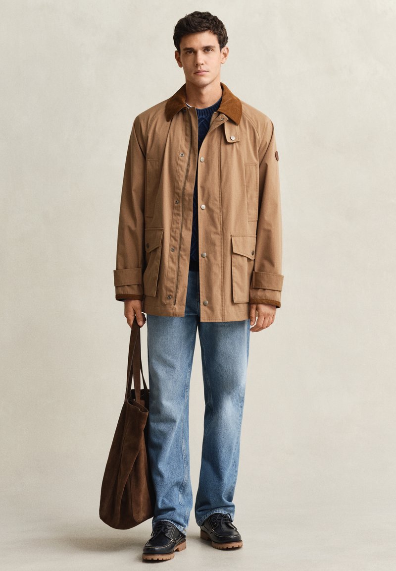 Jeune homme portant une veste beige avec col marron, jean bleu, chaussures noires, tenant un sac fourre-tout marron, debout devant un fond uni.