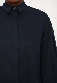 Camicia blu navy con colletto strutturato, chiusura con bottoni al collo e maniche larghe realizzate in tessuto morbido.