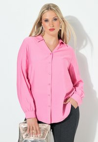 Rosa Langarmbluse mit durchgehender Knopfleiste, weichem Material und Kragen. Getragen mit einer silbernen Clutch und grauen Hosen.