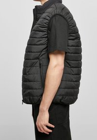 Černá nadýchaná vesta s vysokým límcem, s horizontálním prošíváním, předním zipem a bočními kapsami. Nosená přes černé tričko s krátkým rukávem.