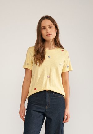 Kvinde med langt brunt hår iført gul kortærmet skjorte med små flerfarvede skaller og mørkeblå højtaljede jeans.