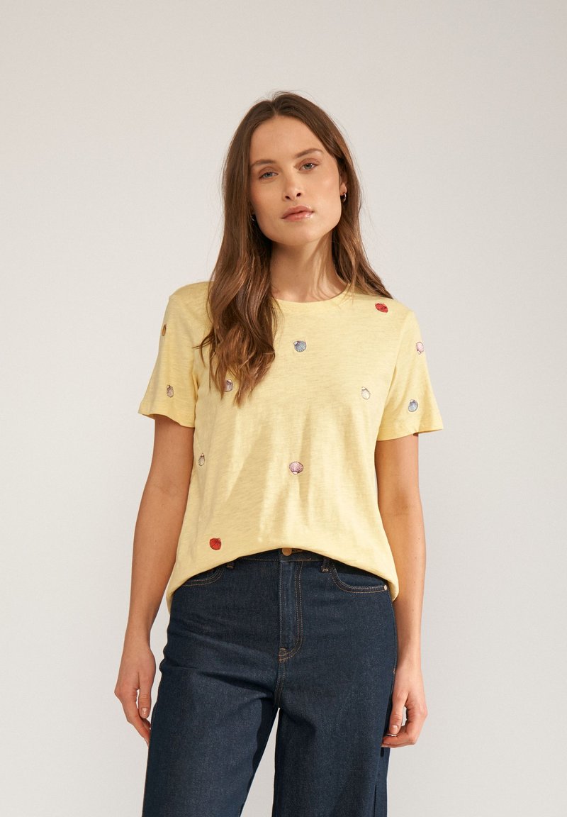 Femme aux longs cheveux bruns portant un t-shirt jaune à manches courtes avec de petites impressions coquillages multicolores et un jean bleu foncé taille haute.