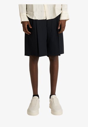 Osoba nosząca czarne plisowane krótkie spodenki do kolan, kremową koszulę w paski z długim rękawem, białe trampki i czarne skarpetki na białym tle.