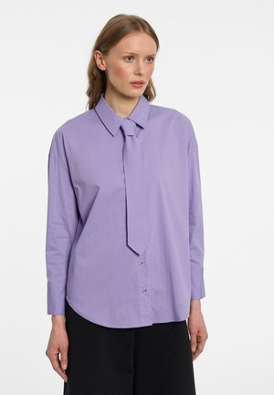 Femme aux longs cheveux châtain clair, portant une chemise lavande à manches longues avec une cravate assortie et un pantalon noir, légèrement tournée vers la droite.