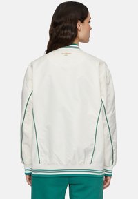 Krämfärgad bomberjacka med gröna detaljer och ränder. Har ett quiltat mönster och ribbade ärmslut. Broderad "Grundad 1911" på baksidan.