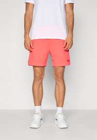Calções de banho corais com um padrão texturizado, apresentando um logótipo preto na perna esquerda. Combinados com uma t-shirt branca que transfere a humidade e ténis brancos.