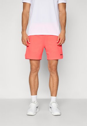 Korallenfarbene Badeshorts mit einem strukturierten Muster, versehen mit einem schwarzen Logo am linken Bein. Kombiniert mit einem weißen feuchtigkeitsregulierenden Shirt und weißen Sneakers.