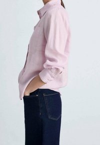 Helles rosa Hemd mit Knopfleiste, einer Brusttasche, aufgerollten Ärmeln und einer lockeren Passform, kombiniert mit dunklen Jeans. Glatte Stoffstruktur.