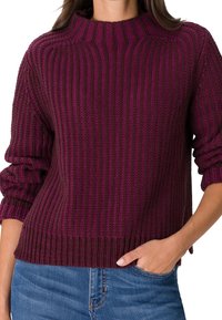 Pull en maille bordeaux avec une texture côtelée, un col montant et des manches roulées. Associé à un jean bleu, mettant en valeur la coupe décontractée.