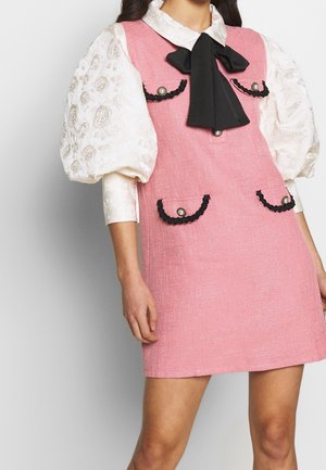 Roze jurk met textuur en een getailleerd ontwerp, versierd met zwarte biezen, parelaccenten op de zakken en een grote zwarte strik bij de hals.