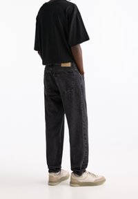 Maglietta nera oversize, pantaloni jogger in denim grigio con polsini elastici e sneakers beige con accenti chiari, vista da dietro.