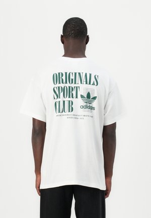 T-shirt en coton blanc avec texte vert indiquant « ORIGINALS SPORT CLUB » et un logo Adidas. Coupe ample, manches courtes, design simple.