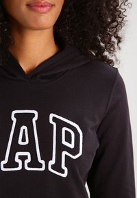 Černá mikina s kapucí, která na přední straně má velké bílé vyšívané logo "AP". Materiál je měkký a kapuce má šňůrku.