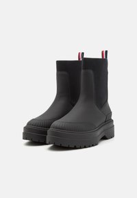 Botas negras de tobillo con exterior de cuero suave, puntera acanalada, suela de goma gruesa y paneles laterales elásticos; cuenta con tiradores en rojo, blanco y azul.