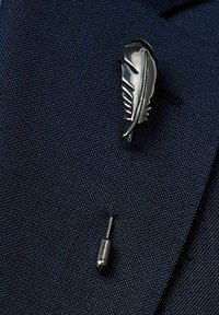 Silverfärgad fjäder brosch, med intrikata detaljer och en polerad finish, fäst på en mörk marinblå kavaj i ett subtilt texturerat tyg.