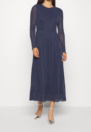 Femme portant une longue robe bleu marine scintillante avec des manches longues transparentes et des chaussures argentées à talons hauts et bride à la cheville, debout sur un fond blanc.