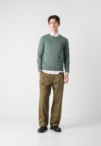 Green knit sweater with a small red logo, layered over a white collared shirt. Brown loose-fit trousers and black shoes complete the outfit.