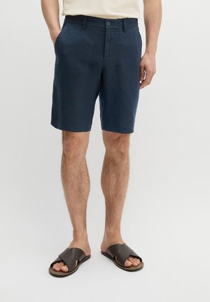 Man draagt marineblauwe knielange korte broek, beige overhemd en donkerbruine sandalen met kruisbanden, staat met één hand in de zak.