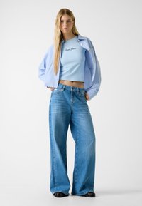 Lichtblauw crop top met "Guess Jeans" tekst, gecombineerd met wijde blauwe denim jeans en zwarte laarzen met hak. Witte achtergrond.
