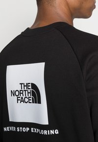 Person som bär en svart sweatshirt med The North Face-logotyp och texten "Never Stop Exploring" på ryggen.