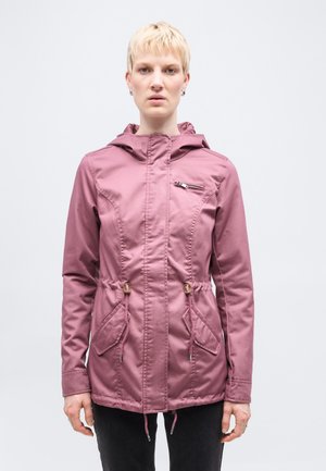 Blond person som har på seg jakke med hette i støvete rosa, med glidelås foran, glidelåslomme på brystet og snøring i midjen, står foran en hvit bakgrunn.