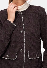 Veste texturée violet foncé avec bordure blanche, dotée d'un col rond, d'une fermeture à boutons et de deux poches avant. Les boutons sont circulaires et métalliques.