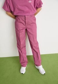 Roze, op maat gemaakte broeken met een rechte snit, gemaakt van gestructureerde stof. Gecombineerd met witte sneakers op een groene vloer.