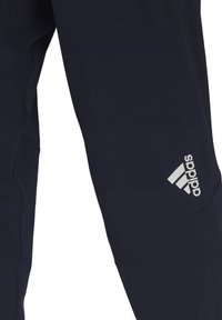 Marinblå idrottsbyxor med slim fit, mjuk textur och en framträdande vit Adidas-logga på nedre benet. Har förstärkta sömmar.