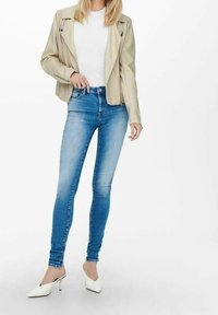 Beige lädermotorcykeljacka över en vit t-shirt, parat med blå slim-fit jeans och vita spetsiga klackar. Släta texturer och åtsittande design.