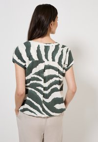Blusa de manga corta con un patrón abstracto de cebra en verde y blanco, hecha de una tela ligera, con un corte relajado y detalle de pliegues en la parte posterior.
