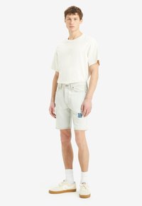 Camiseta blanca de algodón de manga corta, combinada con pantalones cortos de mezclilla claros con un parche azul, complementada con zapatillas beige y calcetines blancos.