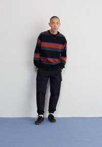 Zwarte gestreepte trui met oranje en blauwe horizontale lijnen, gecombineerd met zwarte cargo-broeken en donkere sneakers, op een effen achtergrond.